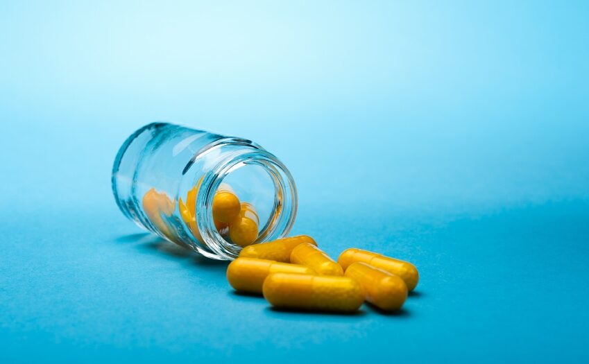 Een glazen pot is omgevallen, gele capsules vallen op een blauw oppervlak. De levendige achtergrond versterkt het contrast tussen de container en de capsules.