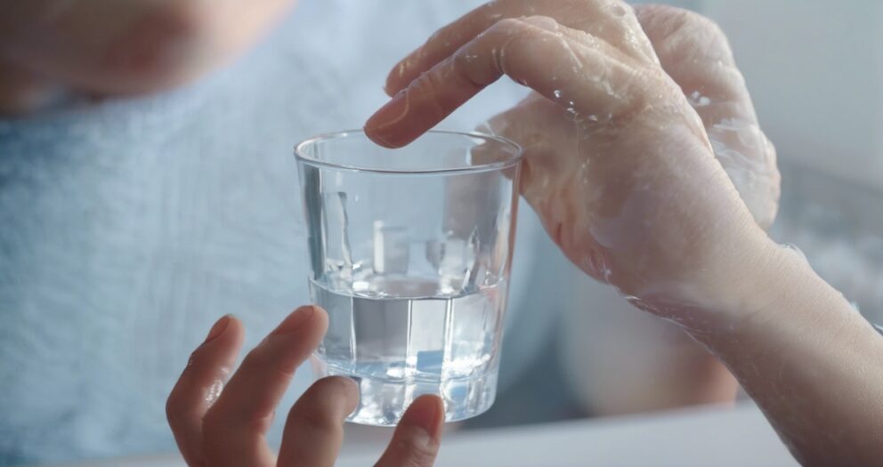 Een close-up van twee handen die een helder glas water vasthouden. De hand links ondersteunt de onderkant van het glas, terwijl de andere hand het van bovenaf vasthoudt. Er zijn waterdruppels zichtbaar op de handen.